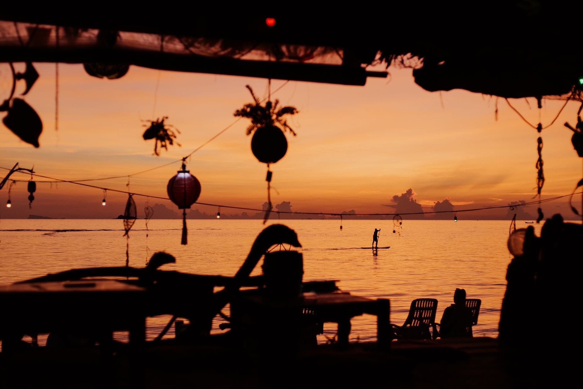Bar na plaży o zachodzie słońca z lampionami i sylwetką paddleboardu